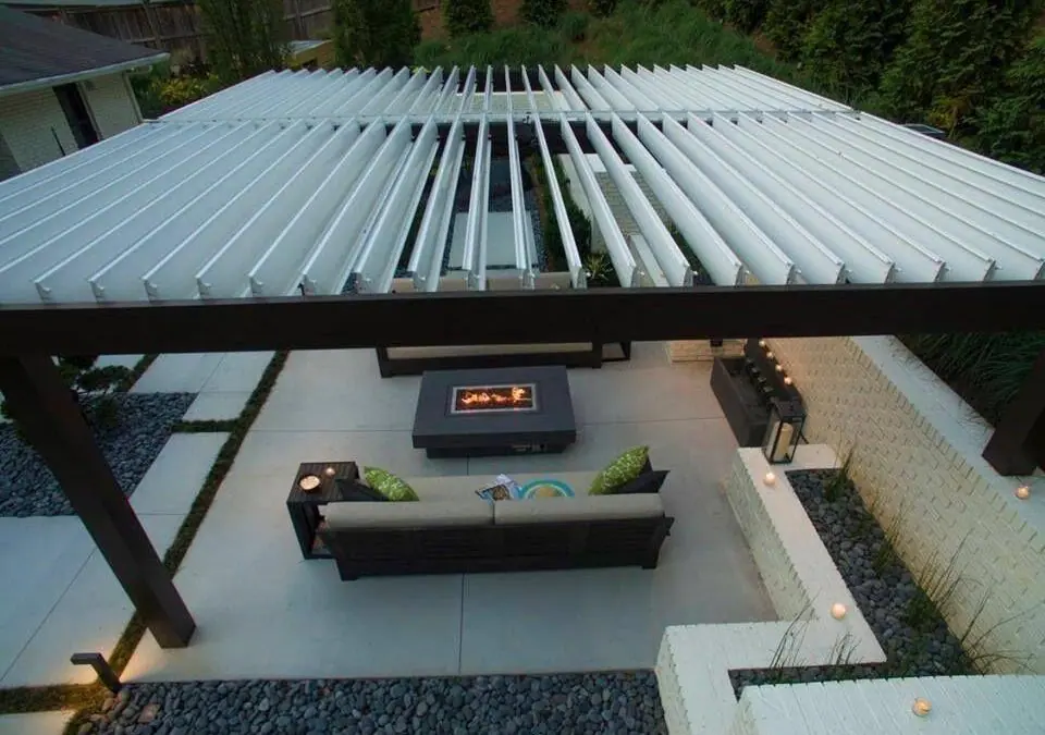 Aerial view of modern backyard patio with white louvered pergola roof and central fire pit surrounded by sectional seating.