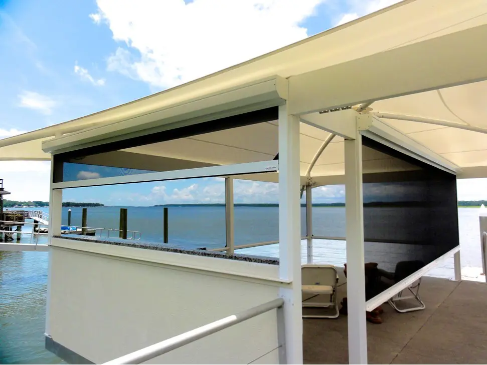 Waterfront patio with white structure and partially lowered black motorized retractable screens, overlooking a calm lake and blue sky
