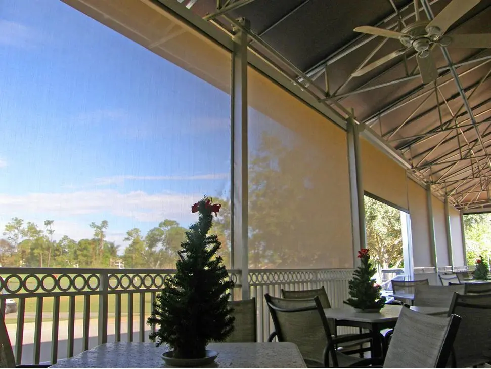 Interior view of a patio with translucent beige roll-down motorized screens, metal frame ceiling, and small decorated Christmas trees on tables.