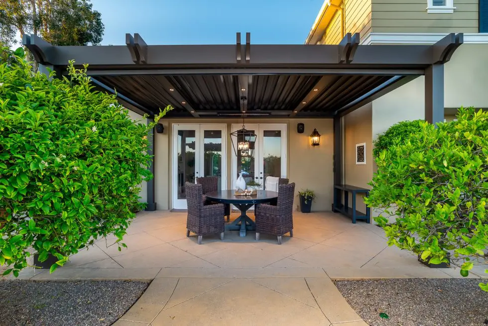 Small cozy patio with wicker furniture and a lantern chandelier beneath a black pergola surrounded by green bushes