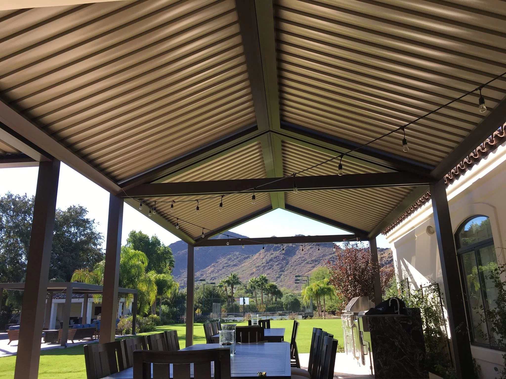 Covered patio with metal gabled roof, string lights, and outdoor dining area overlooking a green yard with mountains in the background.