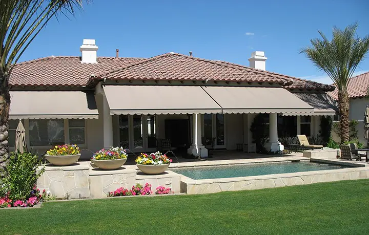 Modern stone patio with a flat pergola roof and a lit water fountain feature, set against a twilight sky and open field.